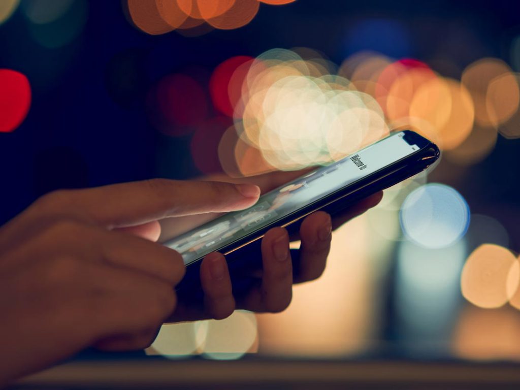 person looking as cell phone screen while out in public at night with lights in the background